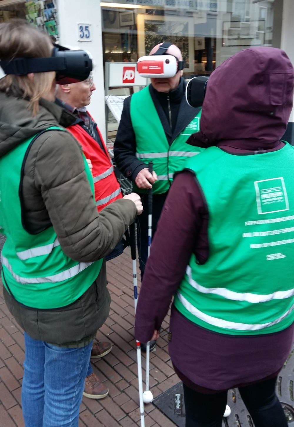 Een groep van 4 mensen die in een kring taan met hun gezicht naar elkaar. Ze dragen waarschuwingshesjes, houden een geleidenstok vast en hebben een VR bril op hun hoofd.