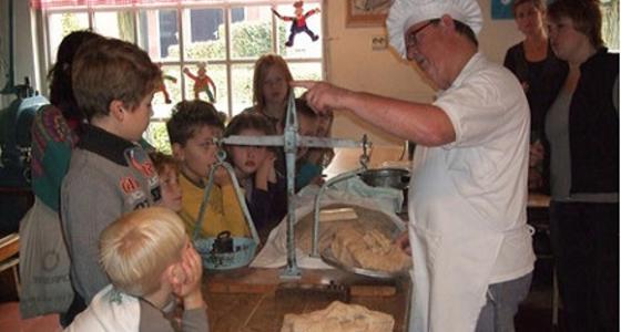 Een groep kinderen staat geïnteresseerd te kijken en luisteren naar een bakker die brood weegt met een ouderwetse weegschaal met gewichten.