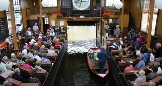 De veilingzaal met in het midden het water waar de boten met goederen doorheen varen en daarboven de veilingklok. Links en rechts de banken met de mensen die willen bieden op de goederen.