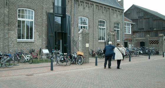 Grijs bakstenen gebouw met hoge ramen en een hoge entreedeur met grote zwarte luiken