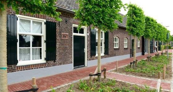 De zijkant van een stenen boerderij met bomen er langs en witte ramen met zwarte luiken in de gevel.