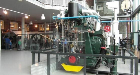 Museumzaal met vide en grote hoge ramen met daarin een grote groene machine met pijpen en stuurwielen.