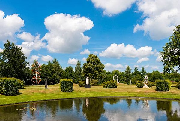 Aan de overkant van het water is een grasveld met struiken en verder naar achter bomen. Op het grasveld staan verschillende beelden en sculpturen.