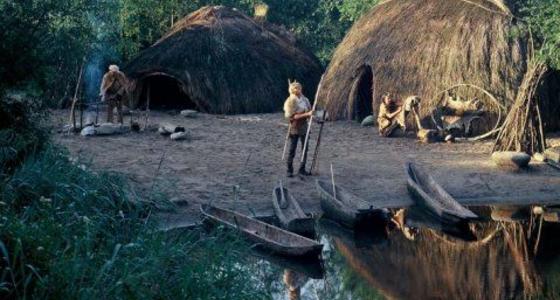 Een historische verbeelding van rieten hutten op een kaal terrein met houten kano's die op de kant liggen en prehistorische mensfiguren.