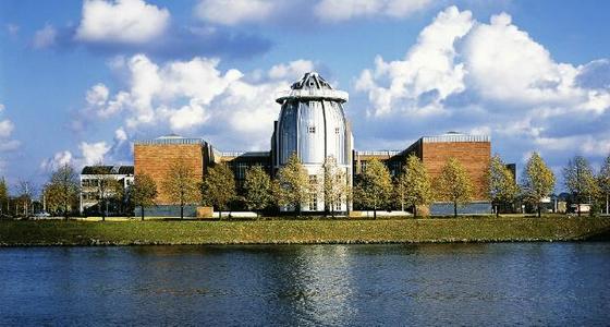 Aan de overkant van het water ligt het museum in de zon. Een grote metalen ronde toren loopt taps toe in een punt, Aan beide zijde wordt de toren vergezeld door rood bakstenen blokken.(gebouwen zonder ramen)