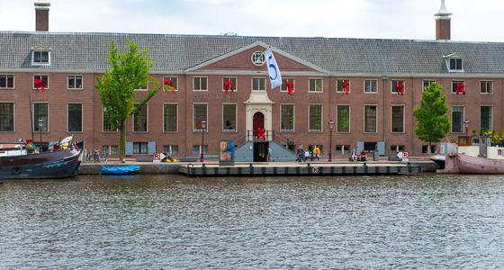 De voorkant van het gebouw de Hermitage aan het water. Het langgerekte stenen gebouw met grijs pannendak is aangekleed met Amsterdamse vlaggen.