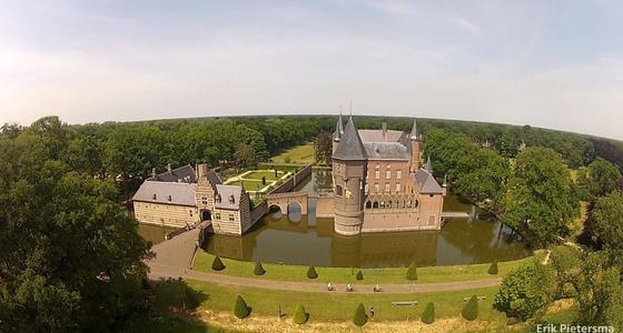 Een luchtfoto van het kasteel met binnen de kasteelmuren een hoog bruin stenen kasteel met zilver grijzen daken, ronde torens en groene tuinen.