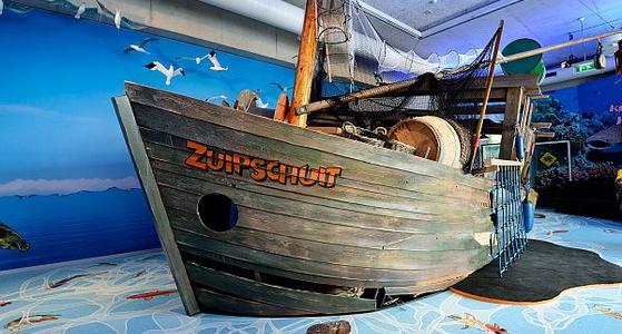 Op de romp van een groot houten schip staat in oranje letters "Zuipschuit", op de boot liggen tonnen en er hangen netten aan de mast. Op de vloer is een zee geschilderd en er hangen meeuwen boven de boot.