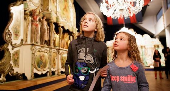 Twee kinderen lopen langs een groot goudkleurig draaiorgel. Ze kijken vol verbazing schuin omhoog naar het orgel met al zijn kleine details.