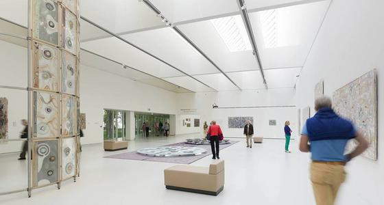 Een grote lichte zaal met witte muren, plafond en vloeren waar kunst tegen de wanden staat en hangt maar ook midden inde ruimte. Een aantal bezoekers bekijkt de kunst in de ruimte.