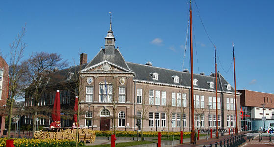 Een statig gebouw van rode bakstenen met een grijs pannen dak. De hoek van het pand heeft een torentje en op het dak staan kleine dakkappellen.