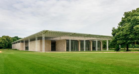 Een modern gelijkvloers gebouw met licht bruine stenen muren en een ingenieus dak voor de beste lichtval naar binnen staat op een groen vlak grasveld