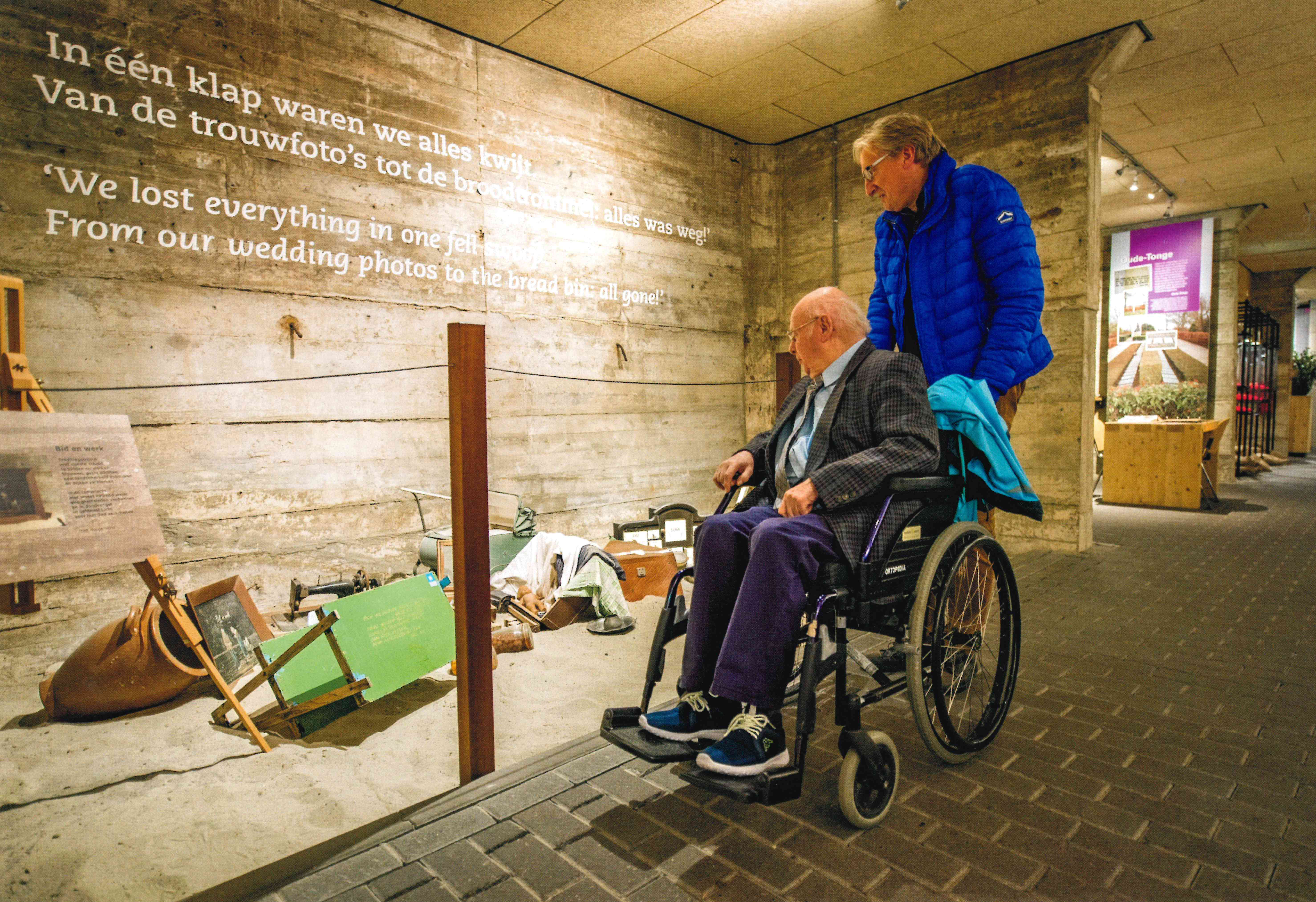 Een man in een rolstoel bekijkt samen met een man die achter de rolstoel staat een deel van de tentoonstelling