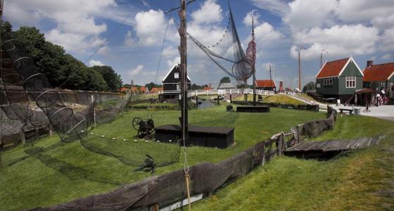 Een omheind grasveld met uitgestalde vissersnetten. op de achtergrond houtenhuisjes en een witte houten gebogen brug.