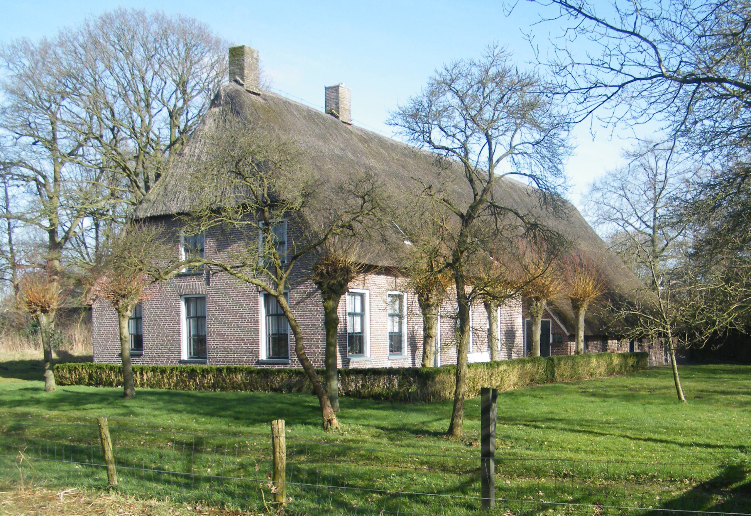 een oude boerderij van lichte bakstenen met rietendak, omringd door een lage haag. Rond de boerderij een grasveld met bomen.