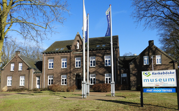 Een statig tweeverdiepingen hoog gebouw van bruine bakstenen en een grijs pannendak. Het gebouw is symetrisch met aan beide zijde van de ingang 2 ramen naast en boven elkaar. Voor het gebouw licht een grasveld met daarin twee hoge vlaggenmasten.