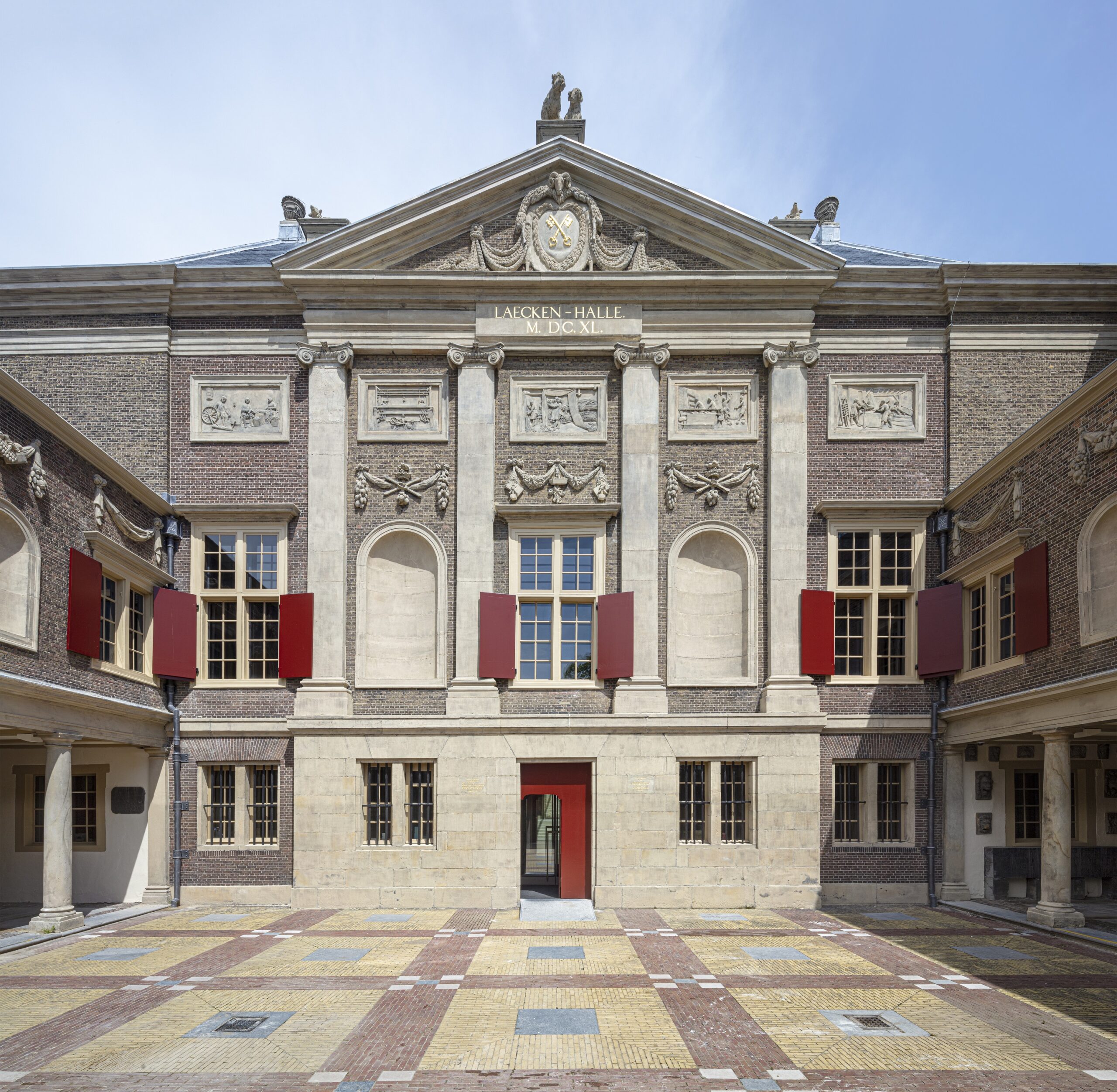 Op een binnenplein met kleurige stenen stat een hoog statig gebouw. Aan weerszijde van het gebouw sluit een galerij met pilaren het plein af. Het licht stenen gebouw wordt opgefleurd door ramen met rode luiken.