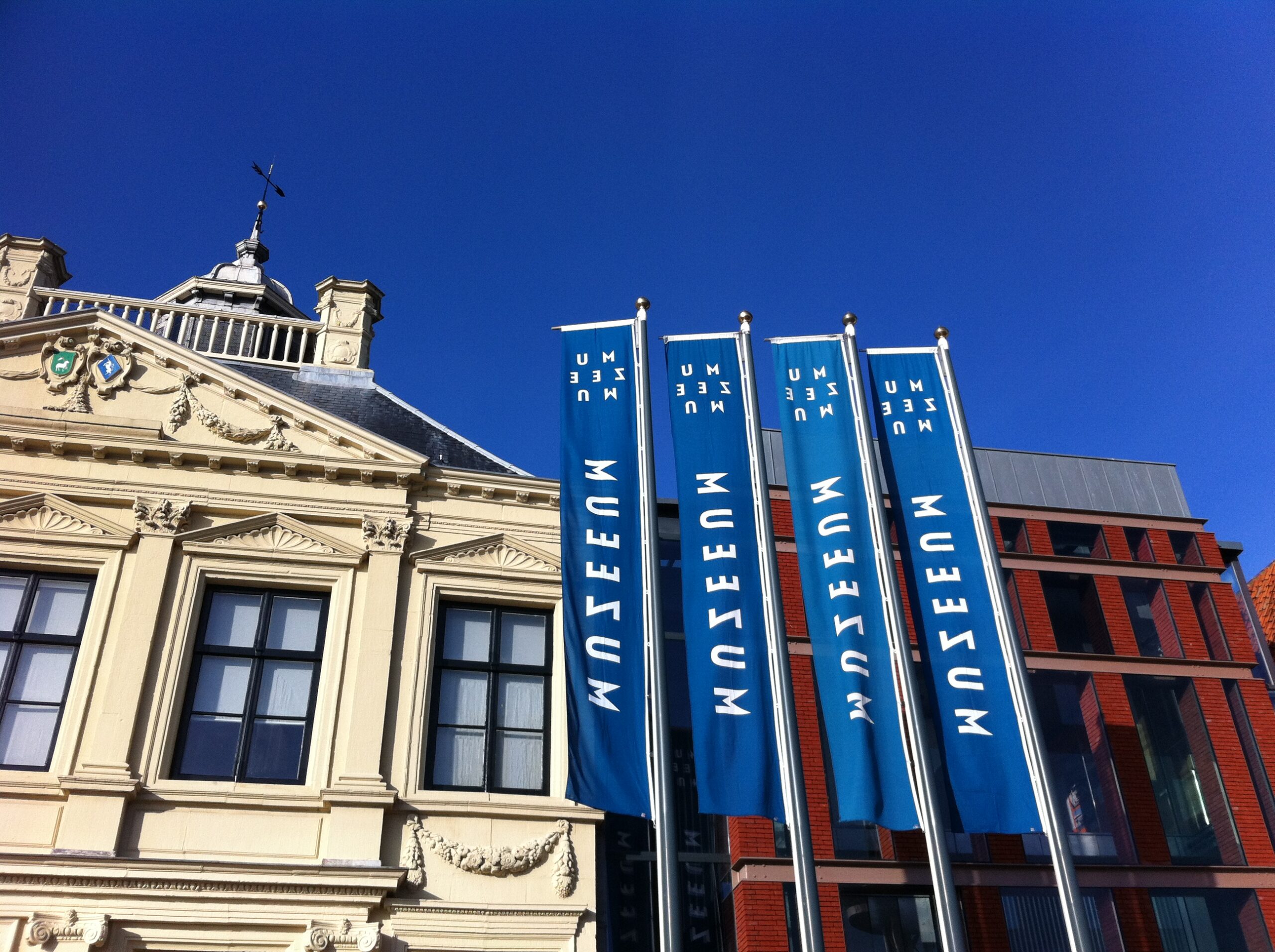 De gevel van een statig cremekleurig gebouw naast een donkerrood modern bakstenen gebouw. Voor de gebouwen 4 vlaggenmasten met blauwe banners met daarop in witte letters de naam van het museum MuZEEum