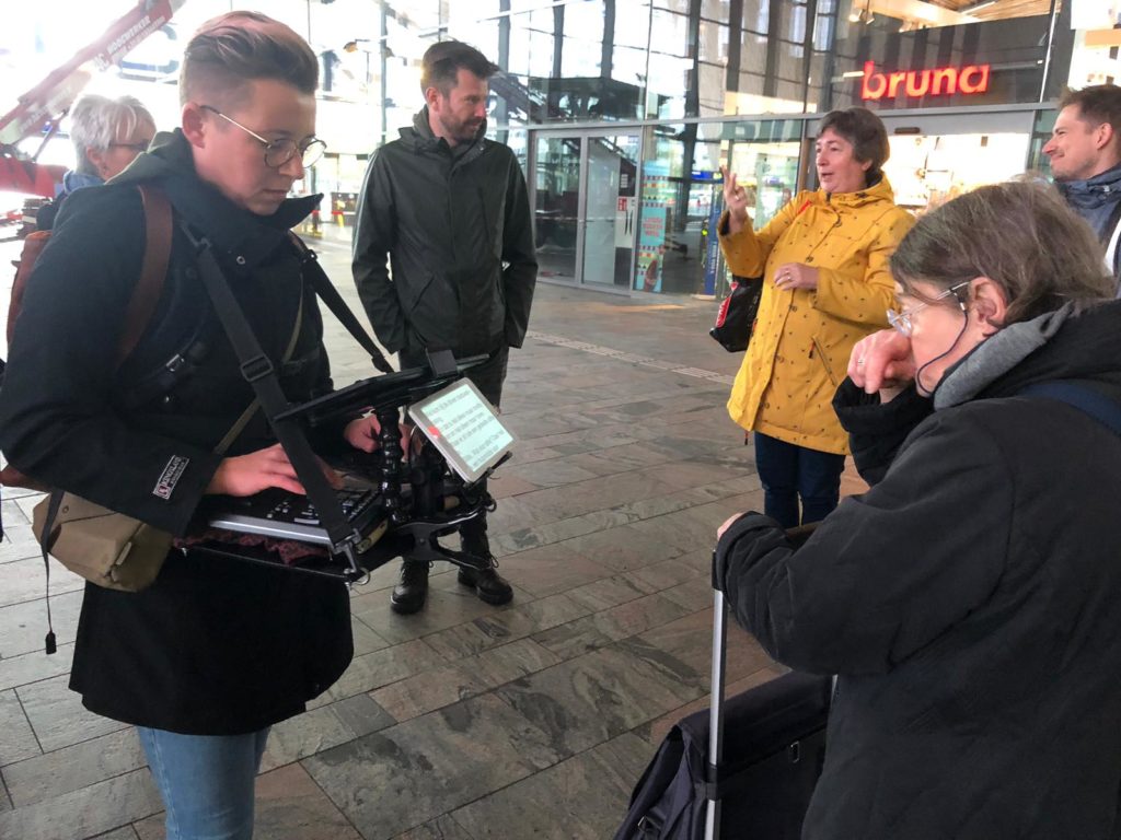 Op de foto staat links een schrijftolk met haar apparatuur, links van haar een man die de gebarentolk is voor de Dove gids. Zij staat daarnaast in een gele jas en gebaart naar de groep. Rechts op de voorgrond staat de slechthorende gids die meeleest op het scherm van de schrijftolk die typt wat er wordt gezegd.