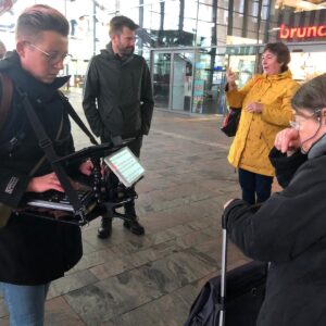 Op de foto staat links een schrijftolk met haar apparatuur, links van haar een man die de gebarentolk is voor de Dove gids. Zij staat daarnaast in een gele jas en gebaart naar de groep. Rechts op de voorgrond staat de slechthorende gids die meeleest op het scherm van de schrijftolk die typt wat er wordt gezegd.