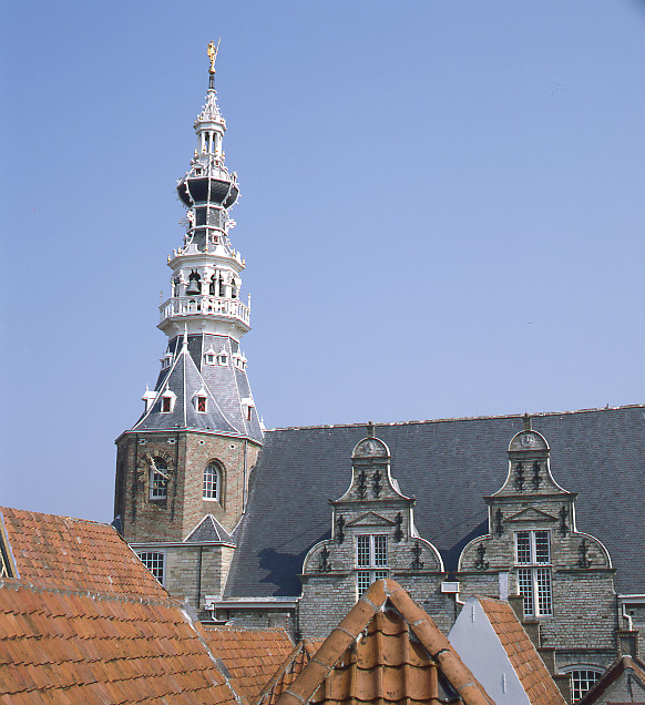 Achter de rode pannendaken, steekt het grijze dak met imposante toren van het stadhuis af tegen de blauwe lucht. Het dak van de toren is opgebouwd ui verschillende lagen met witte balkonnetjes en loopt uit in een punt. De dakkapellen op het dak hebben sierlijke klokgevels.