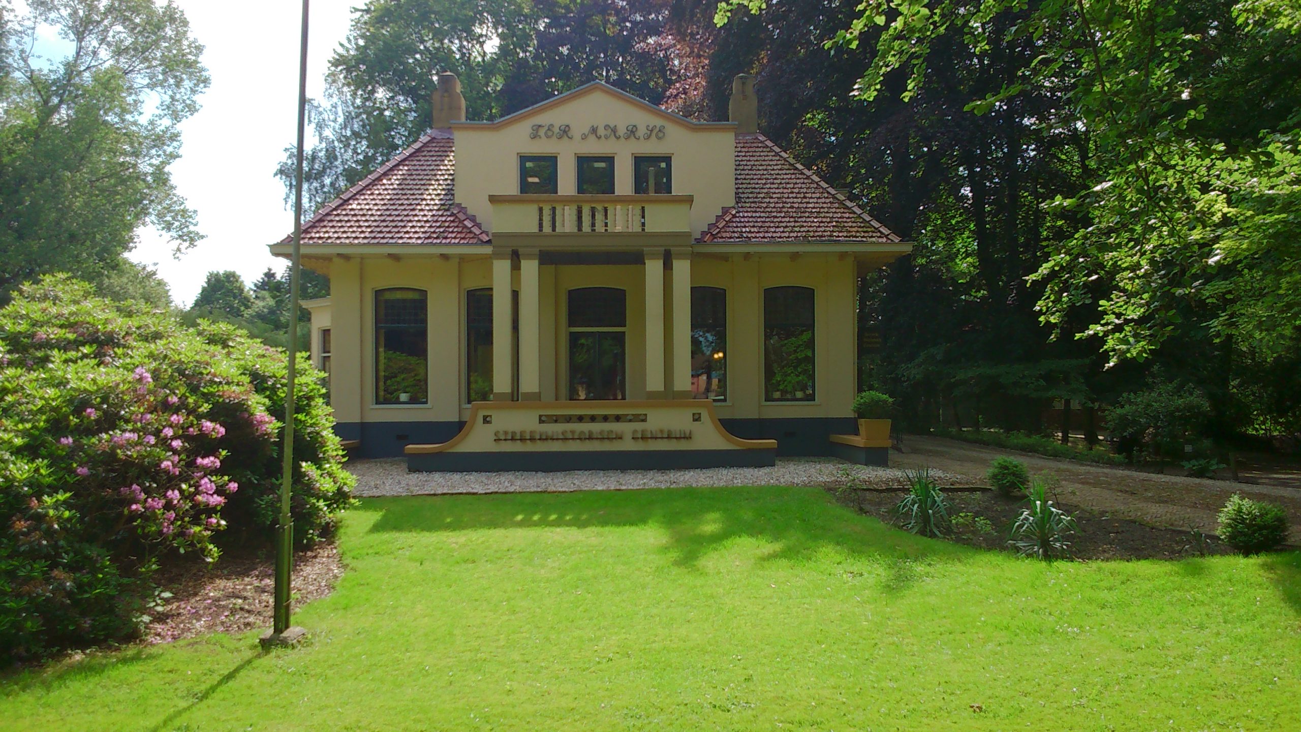 Aan het eind van een groen grasveld, omgeven door bomen, ligt een cremekleurige villa met rood pannendak. Boven de pompeuse ingang met pilaren en een trap ligt een dakkapel met balkon