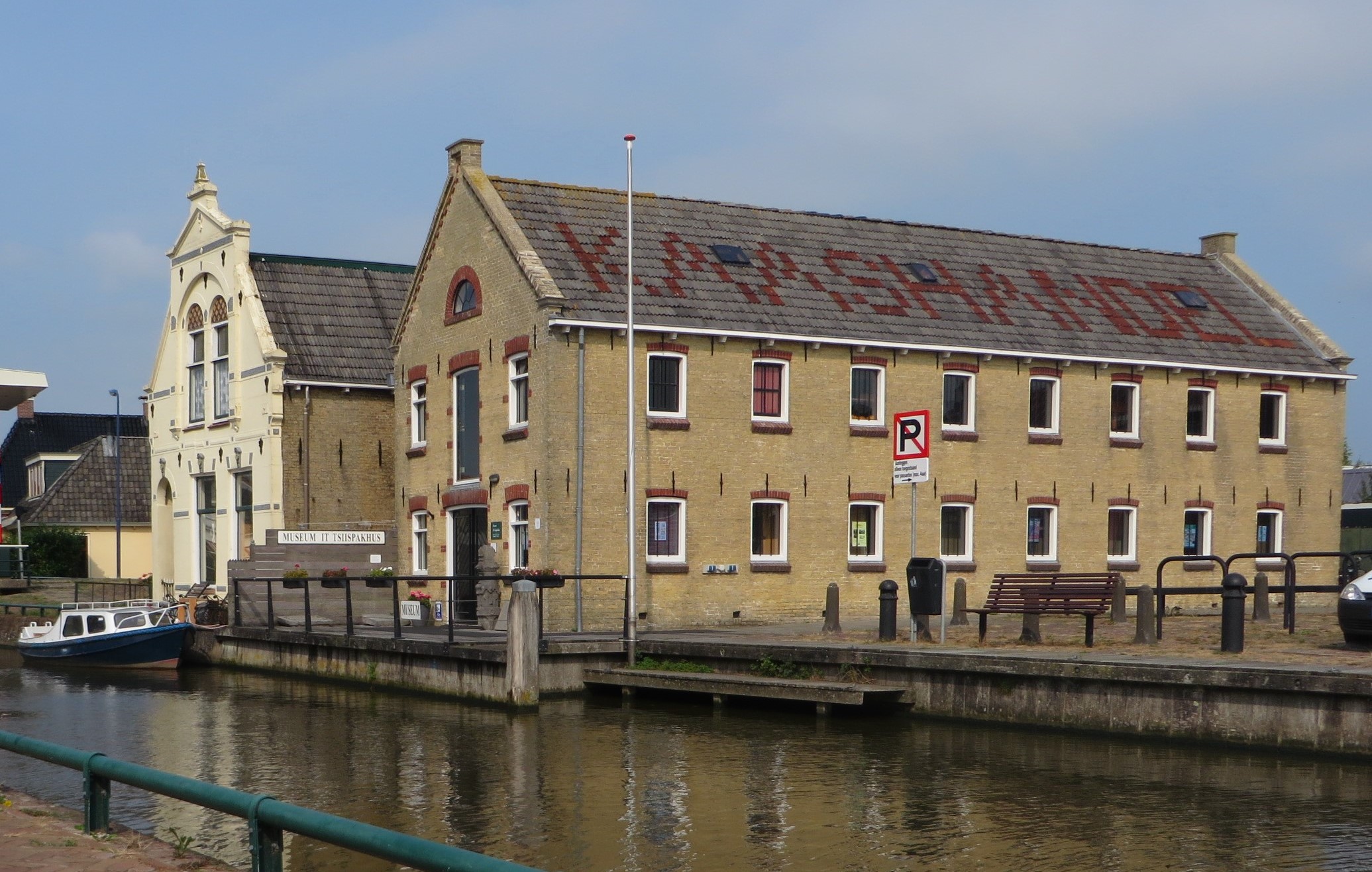 Een zandkleurig stenen gebouw van 2 verdiepingen gelegen aan het water. Het pand heeft een grijs dakpannen waarin rode pannen het woord Kaashandel in is te lezen