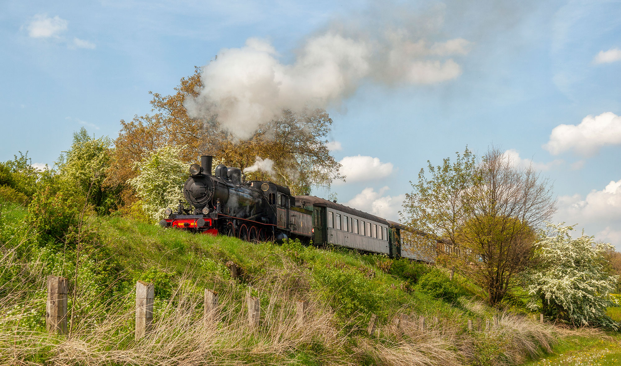 Op een met groen begroeide helling rijd een ouderwets zwarte stoomtrein. Een grote rookpluim komt uit de grote schoorsteen voorop de trein.