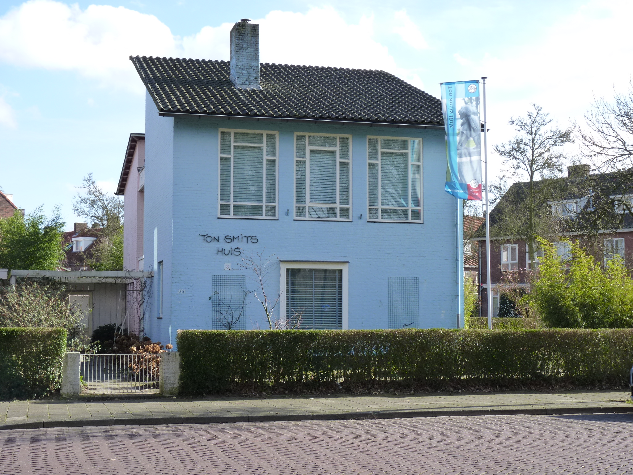 Een klein licht blauw huisje van twee verdiepingen met een groene buxushaag voor de deur. Het huis heeft een grijzen pannendak en het lijkt of het achterste deel van de woning roze is van kleur.