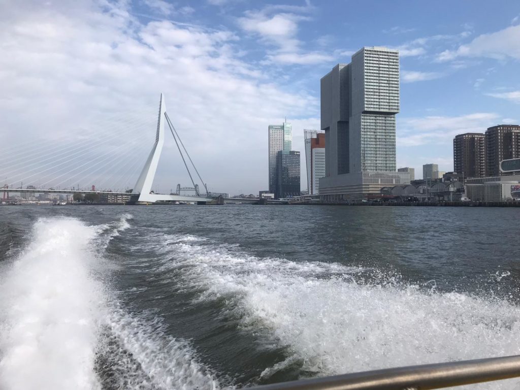 De watertaxi laat een wit schuimend spoor achter in het water met op de horizon de Erasmusbrug en andere inconische gebouwen van Rotterdam