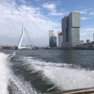 De watertaxi laat een wit schuimend spoor achter in het water met op de horizon de Erasmusbrug en andere inconische gebouwen van Rotterdam