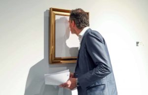 Een man in pak staat voorovergebogen voor een schilderij. Zijn neus raakt het doek bijna. Het schilderij is helemaal wit in een gouden lijst.