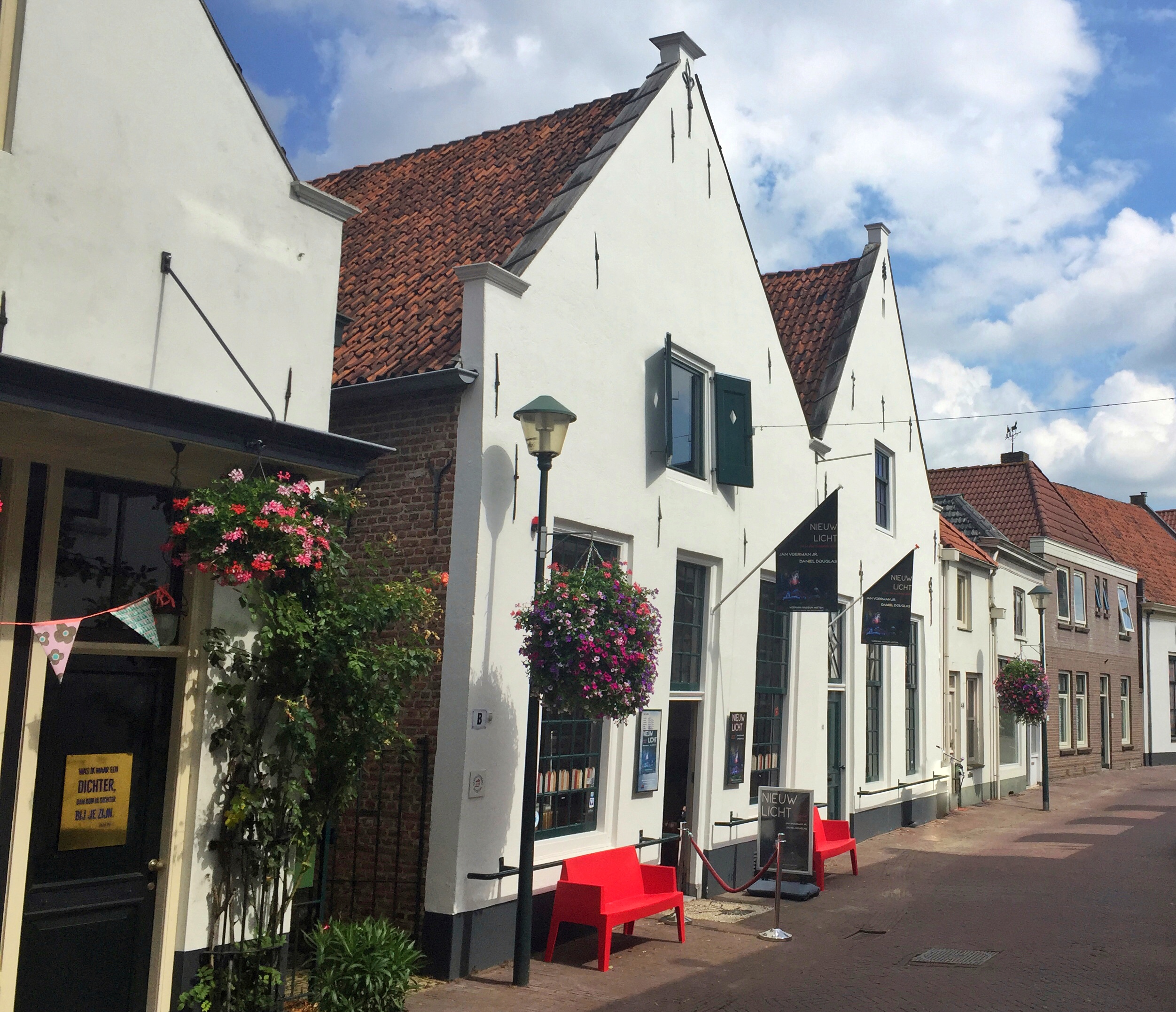 Een wit huis met rode dakpannen, gewoon in een straat. Boven de ingang hangt een vlag en de er staan rode banken onder het raam.