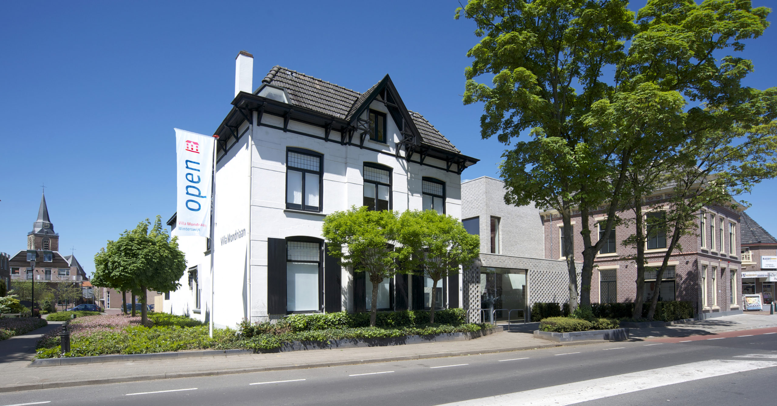 Een witte villa steekt af tegen de strak blauwe lucht. Bij de ingang staan aan beide zijde groene bomen. Het huis ligt aan een asfaltweg met een stoep erlangs.