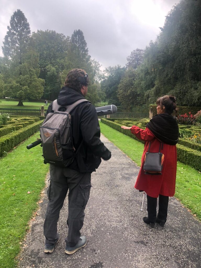 Joan en Edwin staan in het Park en maken opnames voor de audiotour. Joan draagt een rode lange jas met zwarte sjaal en steekt zo mooi af tegen het groen van het gazon en de buxeshagen.