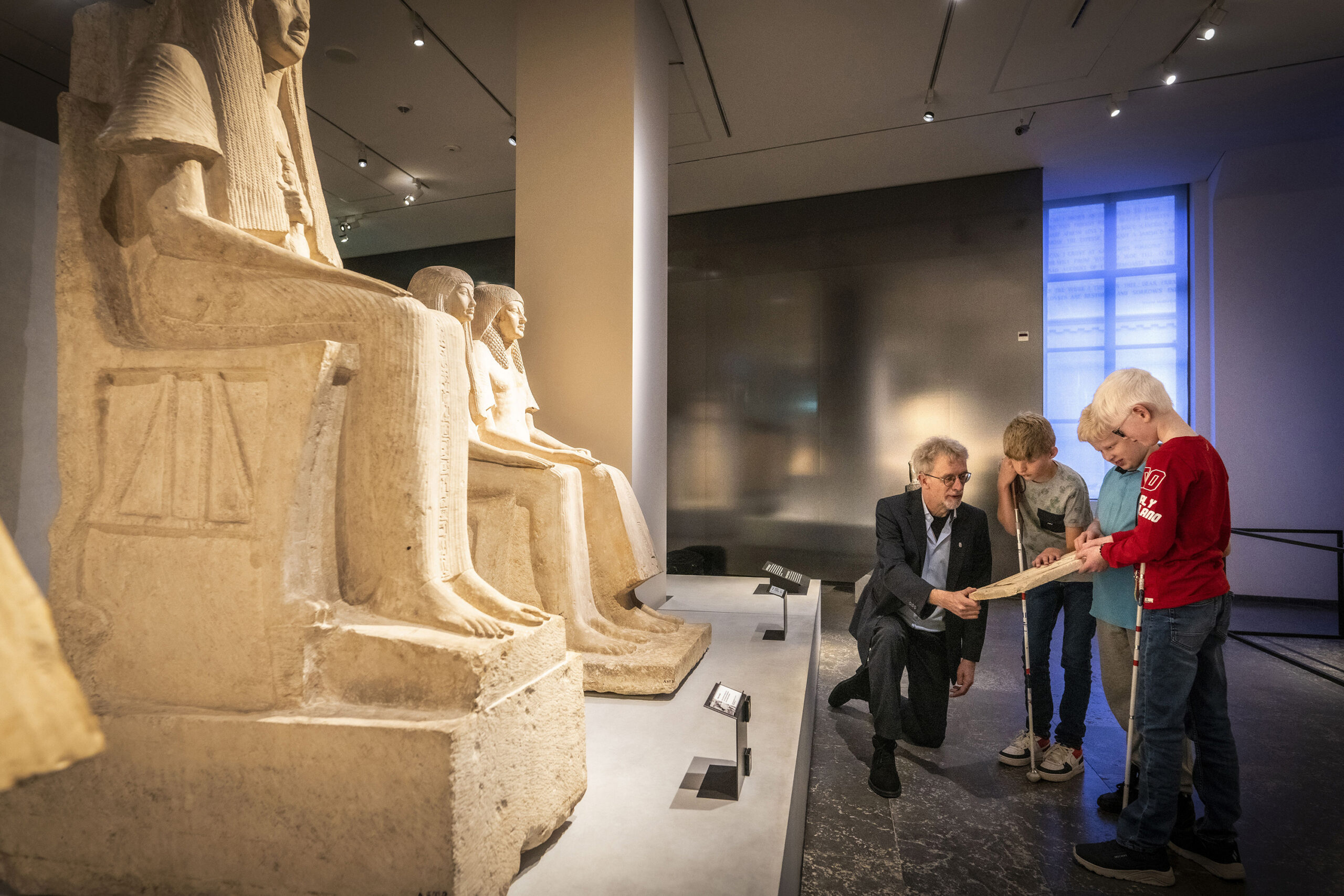 Kinderen staan met een gids in een zaal met grote beelden en voelen aan object