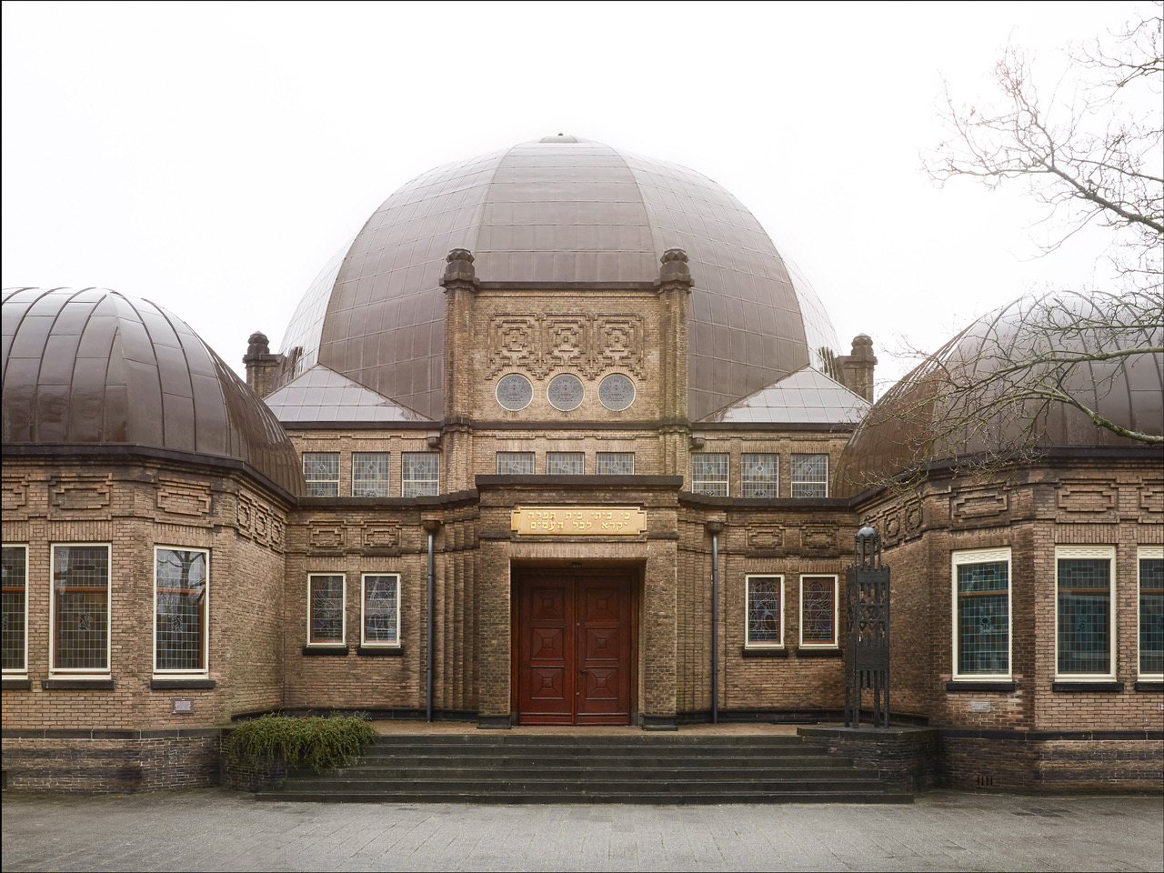 Een foto van de voorzijde van de synagoge met koepelvormig grijs dak. De voorzijde is versierd met gouden schilderingen en heeft een rode brede voordeur.